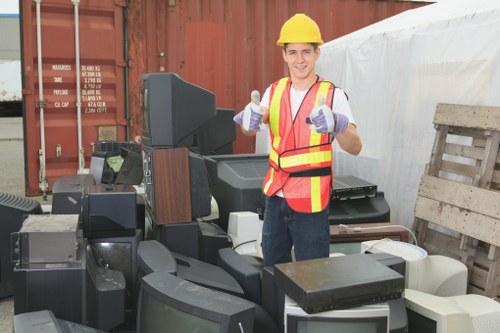 A young male waste management worker wearing a yellow safety helmet, a high-visibility red and yellow safety vest, and purple gloves is standing outdoors on a paved surface near a large collection of discarded electronic devices, including flat-screen monitors, computer towers, and other obsolete appliances. Behind him, a red shipping container is partially visible, suggesting a storage or disposal area, and to his right, a wooden pallet and a white tarp-covered structure can be seen, indicating a typical site for rubbish clearance or bulky waste removal services. The worker is smiling and giving two thumbs up, conveying a positive attitude towards rubbish disposal tasks. The electronic waste appears to be stacked haphazardly, ready for collection or recycling as part of a rubbish clearance service, possibly in an area within Wimbledon or nearby. The environment is well-lit, with natural daylight, and the scene emphasizes the professional and organized approach of Wimbledon House Clearance in managing bulky item and electronic waste removal for residential or commercial clients in the local area.
