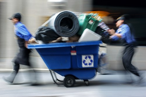 A moving blue recycling bin filled with various discarded items, including black plastic bags, a large black rubber tire, a green plastic container, a white object, and other miscellaneous waste materials, is being pushed along a city street by two workers dressed in blue uniforms and black caps, with their faces blurred due to motion. The background shows an urban environment with blurred buildings and vehicles, indicating an active rubbish removal process typical of services offered in Wimbledon and surrounding areas. The scene captures the dynamic effort involved in clearing waste from residential or commercial properties, reflecting professional rubbish collection services such as those provided by Wimbledon House Clearance. The lighting suggests daytime, with natural light illuminating the scene, and the focus emphasizes the waste materials and the workers engaged in their task.