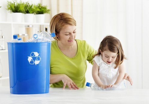 A woman with short blonde hair, wearing a bright lime-green top, is sitting at a table indoors with a young girl who has light brown hair and is dressed in a white top. They are both engaged in a recycling activity, with the woman guiding the girl in handling a clear plastic bottle. To their left, there is a large blue recycling bin with a white recycling symbol on its side, filled with various discarded plastic bottles and containers. Behind them, a white shelf with potted plants and cleaning supplies is visible, and the background features bright, natural light coming through sheer curtains, creating a clean, organized environment that emphasizes waste separation and recycling efforts, which aligns with rubbish management services often offered in the Wimbledon area.