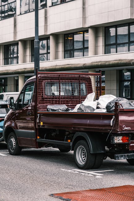 A dark red flatbed truck parked on a city street in front of a modern office building with large glass windows and concrete panels. The truck's cargo area is filled with various types of refuse, inclu