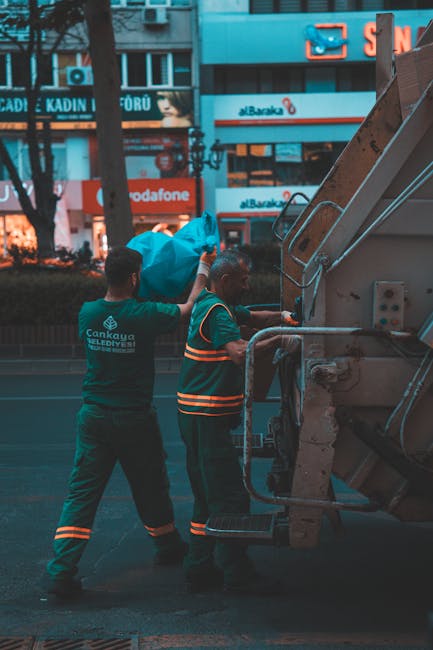 A waste collection worker wearing a high-visibility orange and grey uniform is standing beside a large commercial rubbish truck during dusk or early evening, with a backdrop of dark foliage and an ora